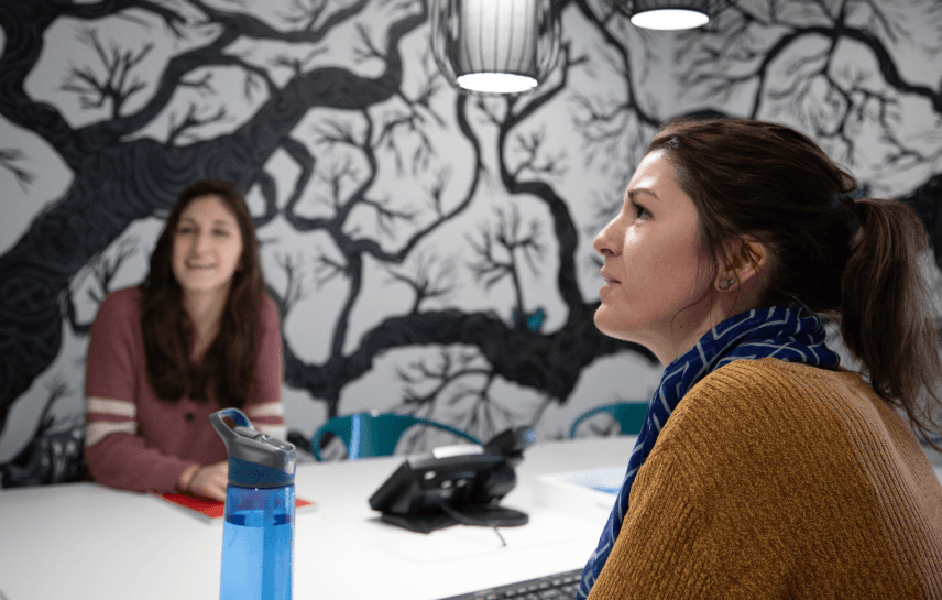 Dos mujeres conversando, una borrosa al fondo.